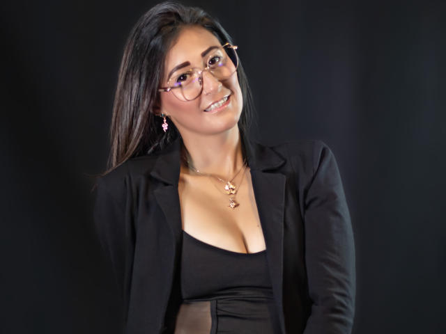 SharissWilliam, con gafas, chaqueta negra y top negro escotado, mostrando los senos, sonriendo, cabello oscuro, aretes rosas y un collar, sobre un fondo negro..