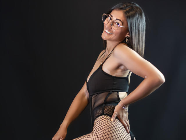 SharissWilliam with glasses, smiling, wearing black mesh lingerie and fishnet stockings, hands on her ass, dark background, light skin, long black hair, gold earrings, sexy pose.