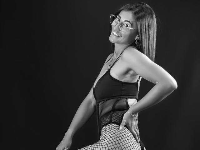 Black-and-white photo of SharissWilliam with glasses, smiling, wearing a sheer black top and fishnet bottoms, showing her ass and small tits, hands on hips, dark background.