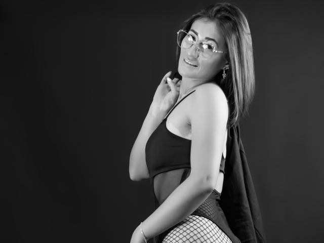 SharissWilliam, de óculos, sorrindo, olhando para trás, vestindo uma blusa preta e calcinhas de rede, mostrando o bumbum, com uma tatuagem no braço. Foto em preto e branco..