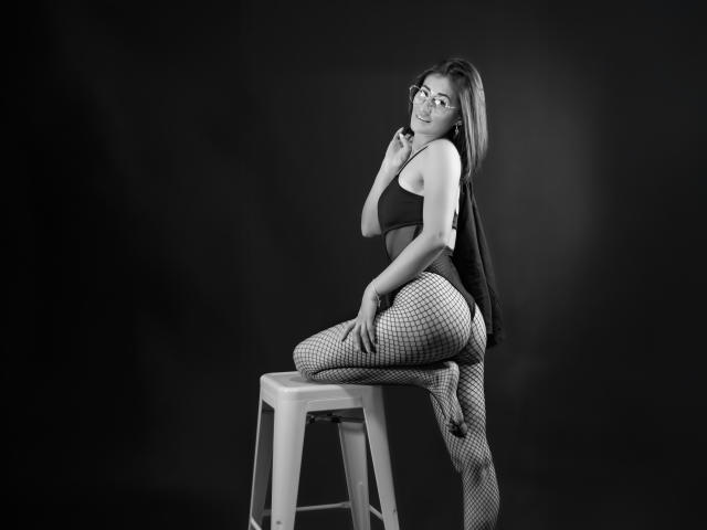 SharissWilliam, avec des lunettes, un débardeur noir, des collants résille et une jupe courte, assise sur un tabouret, le genou droit levé, regardant par-dessus son épaule, souriante, photo en noir et blanc..