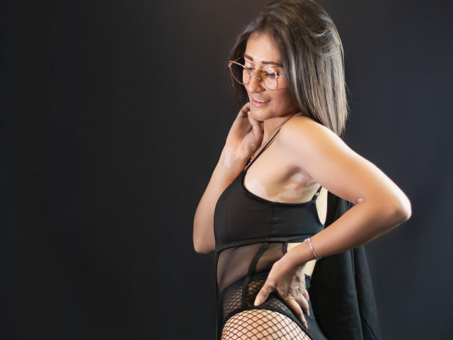 SharissWilliam con ropa interior negra transparente, medias de rejilla y gafas, sonriendo, con la mano en el cuello, fondo negro, perfil lateral, mostrando su trasero y una sonrisa encantadora..