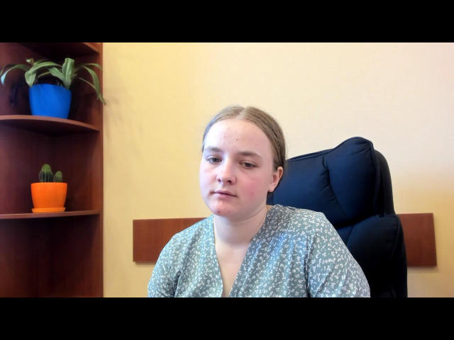 Ellada with pale skin, blonde hair, and small tits, wearing a grey patterned shirt, sitting in a black office chair, looking slightly annoyed, with potted plants on a wooden shelf.