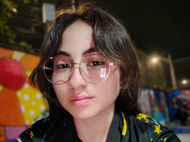 BelleLilith hat helle Haut, dunkles, welliges Haar und pinken Lippenstift. Sie trägt runde, pinke Brillen und eine schwarze Jacke mit gelben Mustern. Ihre Lippen sind leicht geschwollen..