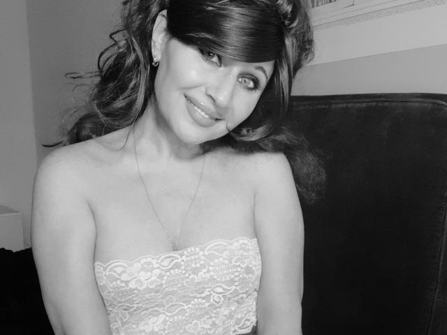 Black and white photo of GingerBett with curly hair, smiling, wearing a lace strapless dress, showing her tits, necklace, and earrings, with dark background.