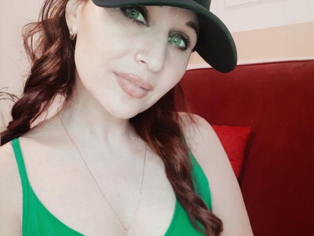 GingerBett with green eyes, wearing a green tank top, black cap, and a necklace, showing her tits, has wavy brown hair, and a slight smile.