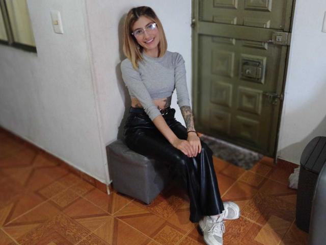 Kaiix, avec des cheveux blonds, des lunettes, un débardeur gris, un pantalon en cuir noir, des baskets blanches, assise sur un pouf gris, avec un tatouage sur le bras gauche, sur un sol en bois, souriant..