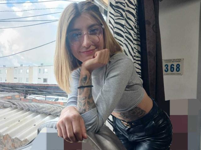 Kaiix leunt tegen een leuning, draagt een bril, een grijze crop top en zwarte leren broek, heeft tatoeages op haar arm, en er hangt een gordijn met een zebrapatroon achter haar..