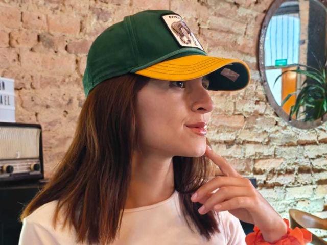 CataleyaSuun con capelli castani, cappello verde e giallo, camicia bianca, dito vicino alle labbra, muro di mattoni sullo sfondo, sguardo di lato, sorriso dolce, stile casual..