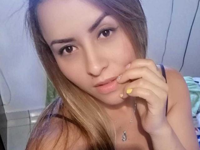 Esposa Nicole, com cabelo castanho claro, olhos castanhos e esmalte de unha amarelo, tocando os lábios com o dedo, vestindo uma blusa preta com alças e um colar com um pequeno pingente..