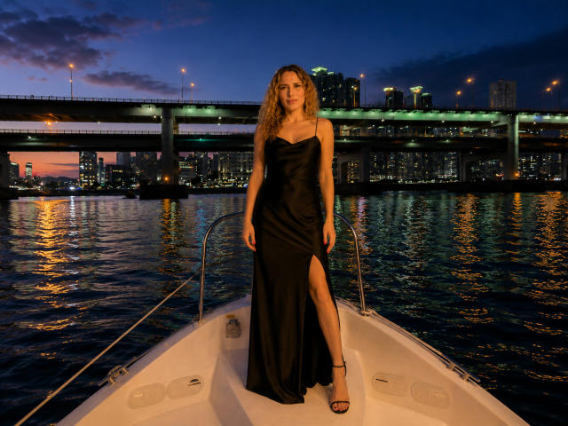 TessaDaybreak with long blonde hair, wearing a black, strapless, high-slit dress and black heels, standing on a boat at night, city skyline and bridge in the background.