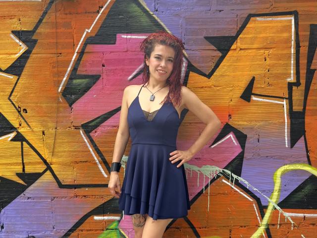ArielleBrooks with red hair, wearing a navy blue dress with lace trim, standing against colorful graffiti, smiling, hand on hip, visible tattoo on left thigh.