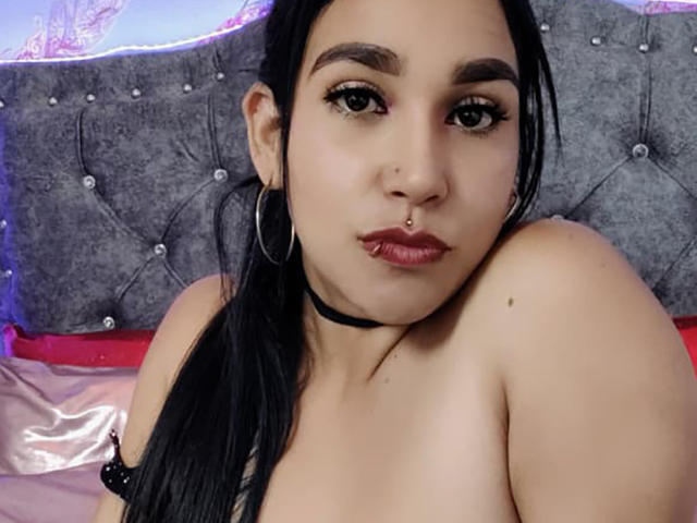 EdenIvyLm with black hair, hoop earrings, choker, nose ring, and pouty lips, showing bare shoulders, looking at camera, sitting on bed with gray tufted headboard.