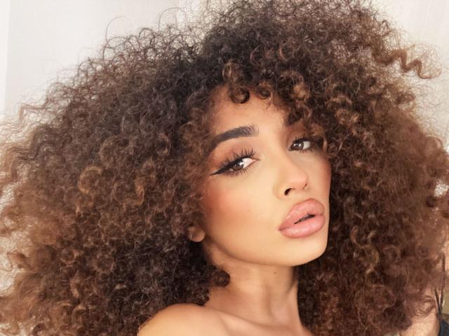 ChloeBlowie con un grande afro riccio marrone, labbra piene rosa, sopracciglia scure e folte e lunghe ciglia, che guarda leggermente di lato con un'espressione seducente..