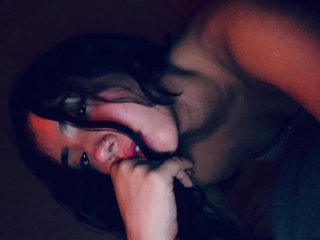 AbrilKoss with dark hair, nose ring, and red lips, topless, biting her right arm, showing her bare shoulders and soft skin, in a dimly lit, intimate setting.