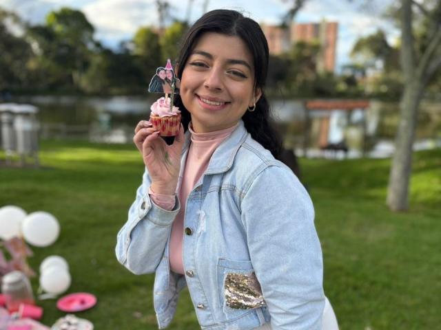AnntonellaWatson with long dark hair, smiling, holding a cupcake with pink frosting and sprinkles, wearing a pink turtleneck and light blue denim jacket, standing outdoors.