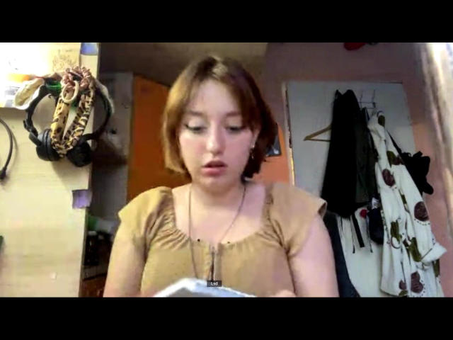 AlaneRodrigo has short brown hair, light skin, and small tits, wearing a tan shirt, looking down with a slightly open mouth, and a necklace. She's in a cluttered room.