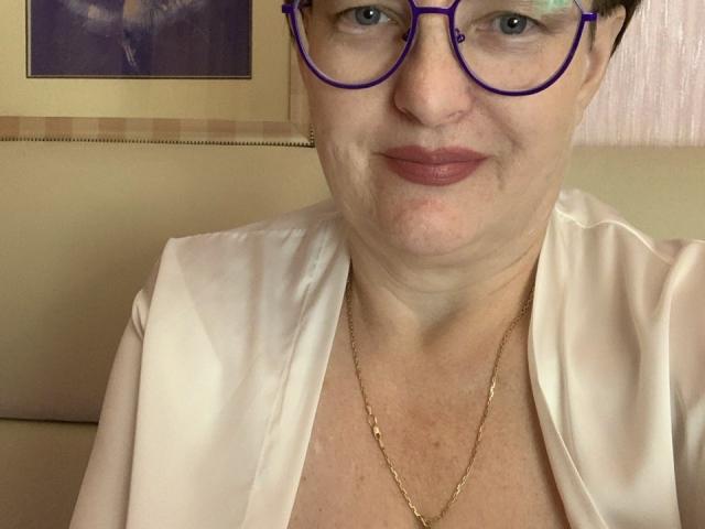 Margie Evans, con gafas moradas, blusa blanca, collar dorado y lápiz labial rosa, mostrando su rostro y parte superior del pecho. Tiene el cabello corto y negro y una ligera sonrisa..