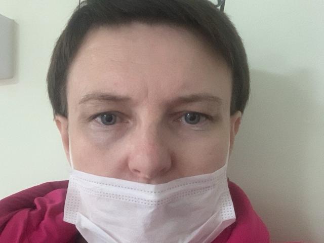 MargieEvans with short brown hair, blue eyes, and a white mask covering her nose and mouth, wearing a red hoodie. Her face shows a slight frown.