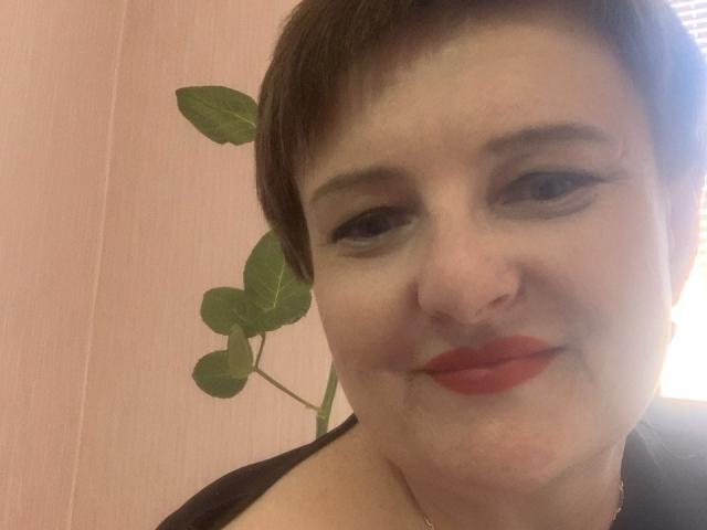 Margie Evans, avec ses courts cheveux bruns, son rouge à lèvres rouge et son anneau de nez, sourit à l'appareil photo. Une plante verte à feuilles est visible en arrière-plan. Le mur est d'une douce couleur rose. La lumière est douce..