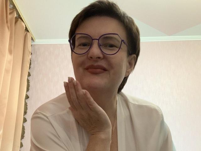 MargieEvans with short brown hair, purple glasses, and a light pink shirt, smiling gently with her hand near her face, showing a subtle, relaxed vibe.
