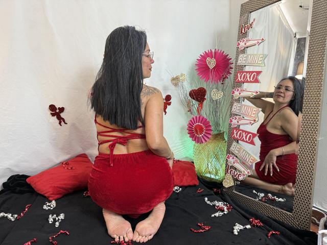 LailaCastillo, assise sur un lit noir, les genoux repliés, portant une robe rouge en velours avec un dos croisé, se regarde dans le miroir. Elle a de longs cheveux noirs et un tatouage sur l'épaule droite. Son reflet est visible..