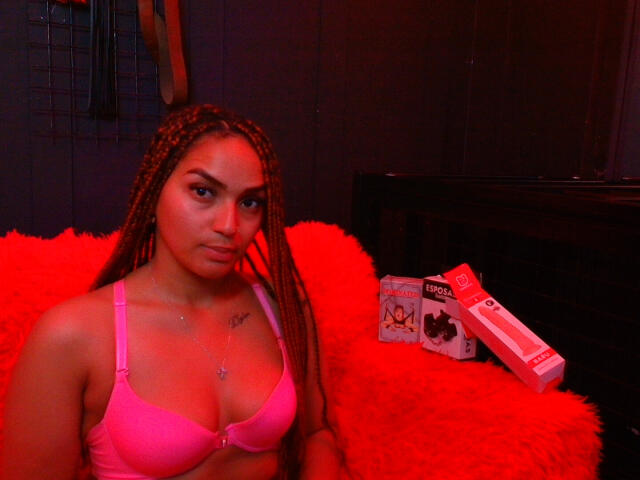 BrianaPratt with braided brown hair, pink bra, and red fuzzy blanket, sitting with a box of L'Oréal condoms on a black bed, looking at the camera.