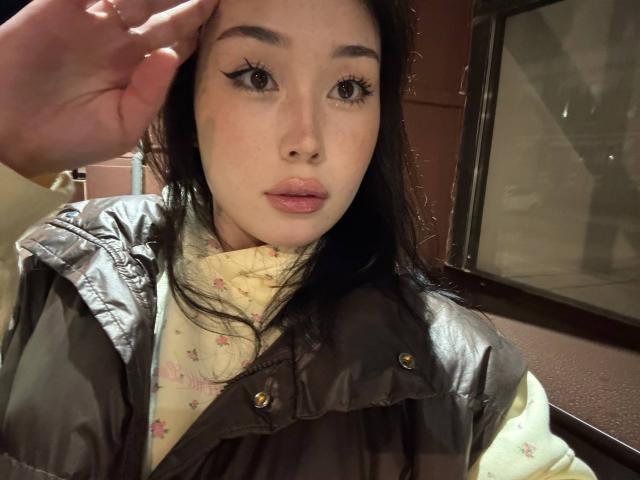 LorenzaMccaughan with dark hair, pale skin, black eyeliner, and pink lips, wearing a gray jacket over a yellow floral shirt, touching her head.
