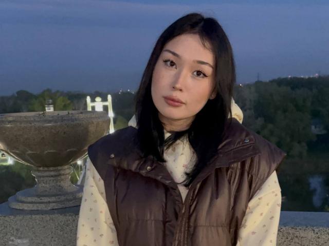 LorenzaMccaughan with black hair, light skin, brown eyes, wearing a puffy brown vest and patterned shirt, standing near a stone fountain at dusk.