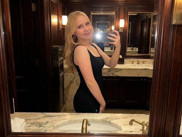 Cayla Henby, con cabello rubio, vestida con un vestido negro, mostrando su trasero en un selfie frente a un espejo, sosteniendo un teléfono, en un lujoso baño con encimeras de mármol..