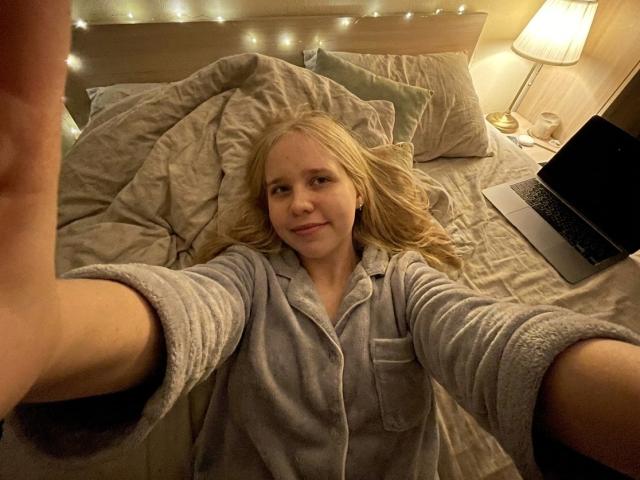 CaylaHenby sdraiata su un letto, indossa un accappatoio grigio, con lunghi capelli biondi, sta scattando un selfie, un laptop è a destra, luce soffusa, sorridente, letto leggermente disordinato..