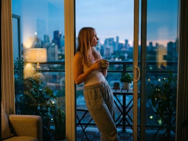 Cayla Henby mit langen, blonden Haaren, die ein weißes Tanktop und graue Jogginghosen trägt, steht an einem Glasbalkon, hält eine Tasse in der Hand und blickt in den Abendhimmel auf eine Stadtansicht..