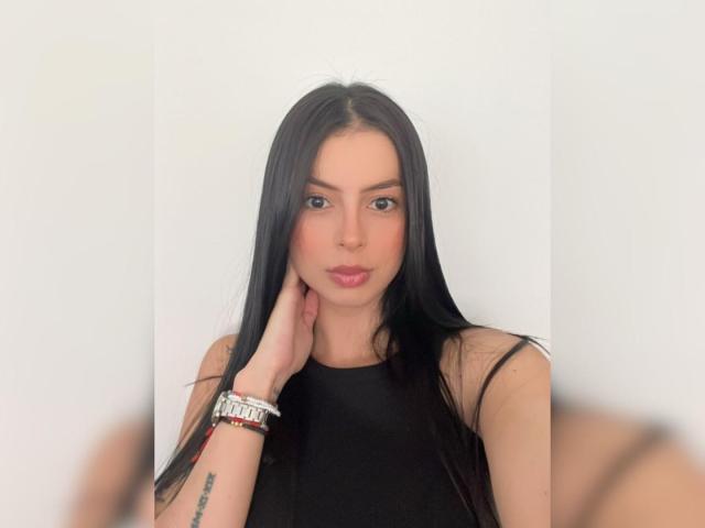 IsabellaJames with long black hair, wearing a black sleeveless top, looking at the camera, pink lips, hand on cheek, silver bracelet, and tattoo on her right wrist.