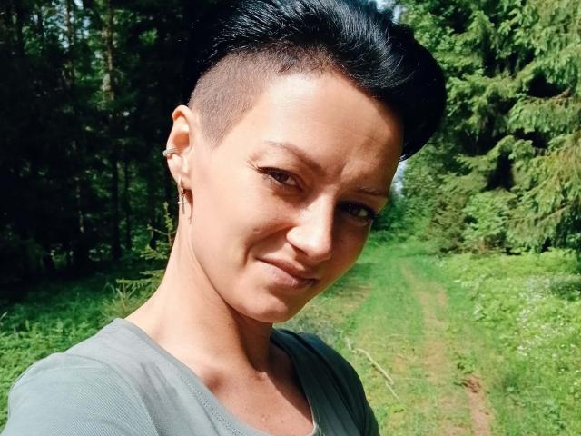 EhidnaLowq hat kurze, schwarze Haare, eine rasiert Seite und ein kleines Ohrpiercing, trägt ein graues Hemd, lächelt leicht und steht in einem grünen Wald mit einem schmutzigen Pfad..