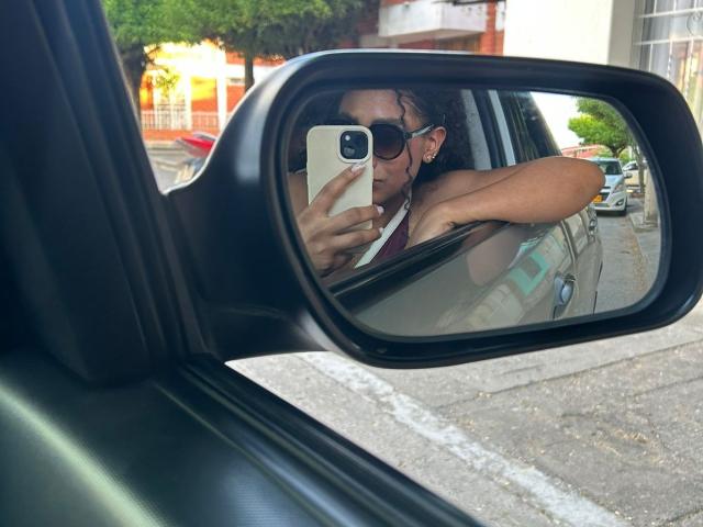 NatalyBlonder mit lockigem, braunem Haar, die eine Sonnenbrille und ein pinkes Tanktop trägt, macht ein Selfie im Rückspiegel des Autos und zeigt dabei ihre freie Schulter und ihre kleinen Brüste..