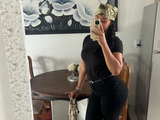NatalyBlonder mit großem Hintern, trägt ein schwarzes T-Shirt, schwarze Hose, einen goldenen Kopftuch und hält ein Telefon, macht ein Spiegel-Selfie, steht neben einem Holztisch und hält eine Tasche mit Leopardenmuster..