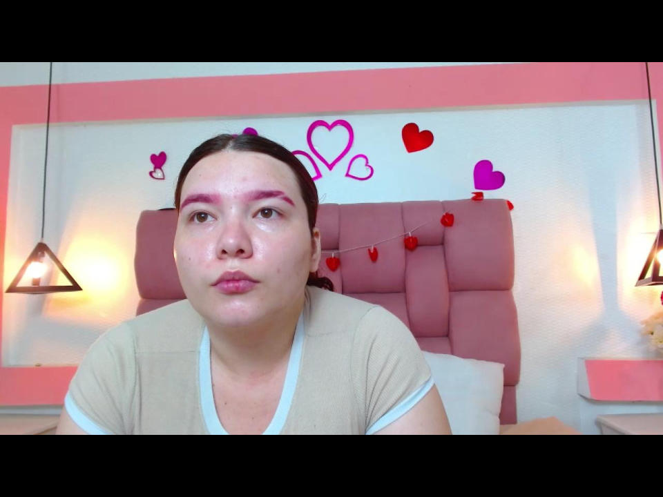 KattyVenus, avec un maquillage rose pour les yeux, des lèvres gonflées et une chemise beige, est assise devant une tête de lit rose ornée de cœurs, et regarde directement l'appareil photo. Des guirlandes lumineuses rouges sont au-dessus de sa tête..