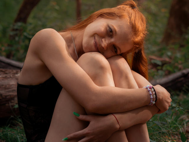 FoxyyMolly with red hair, hugging knees, smiling, wearing black lingerie, green nails, white and purple bracelet, sitting in forest, light brown skin, small tits, cute.