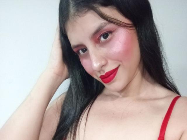 MadissonFontaine, com batom vermelho, sombra rosa e rosto brilhante, sorrindo para a câmera, com a mão no cabelo preto, usando uma alça vermelha, mostrando os ombros nus, charmosa e sexy..