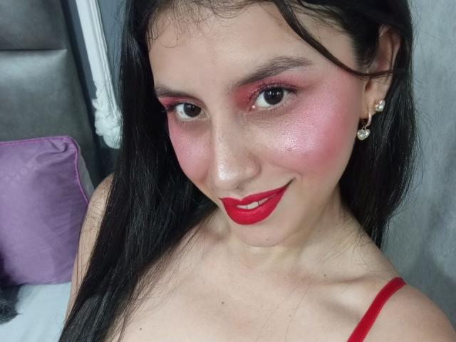 Madisson Fontaine, com bochechas rosadas, batom vermelho e delineador vermelho, sorrindo para a câmera. Ela tem cabelos longos e pretos, usa brincos e está vestindo uma blusa vermelha com alças finas..