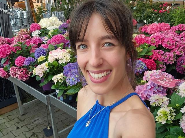 LaylavonHohensee-hot with brown hair, blue eyes, and a smile, wearing a blue halter top, standing in front of colorful hydrangeas. She has a cute face and small tits.