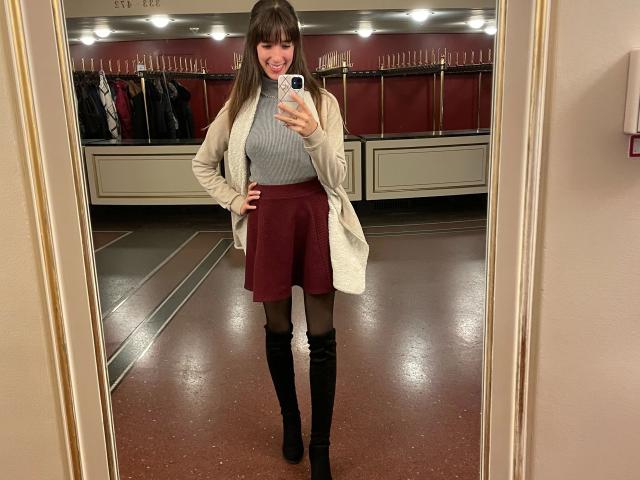 LaylavonHohensee-hot in a mirror selfie, wearing a gray turtleneck, white coat, high-waisted red skirt, black stockings, and black knee-high boots, smiling with hands on hips.
