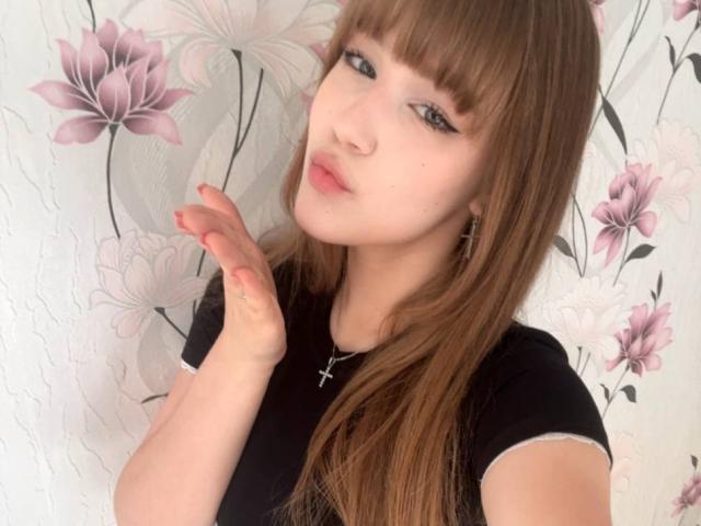 CorinaMakinster has light skin, long brown hair with bangs, wearing a black shirt, and a cross necklace, her right hand up, looking at the camera with pink lips.