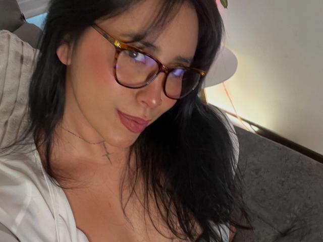 ElizabethMacy with glasses, black hair, pink lipstick, small cross tattoo, and a white shirt, showing her tits slightly. She's looking at the camera with a subtle smile.