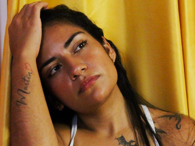EmmaSteele with dark hair, brown skin, and tattoos on her arm and chest, wearing a white tank top, resting her head against yellow curtains, looking slightly pouty.