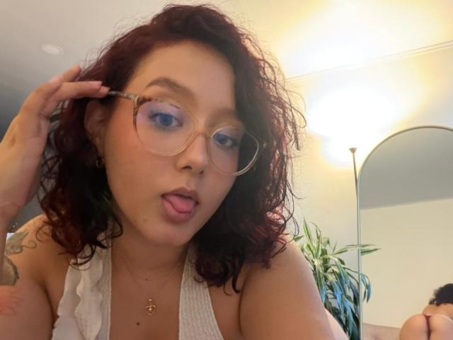 Sophia Browm hat lockiges, rotes Haar, blauen Lidschatten und steckt ihre Zunge heraus. Sie trägt eine Brille, ein weißes Oberteil mit Rüschen und eine goldene Halskette. Ihre Hand ist in ihrem Haar..