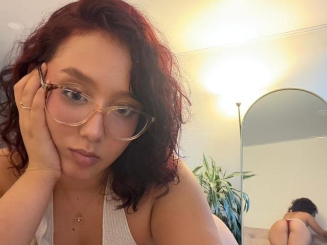 SophiaBrowm with curly red hair, glasses, white tank top, hand on cheek, looking at camera, mirror reflecting her bare ass, plant in background, warm light, slightly pouty expression.