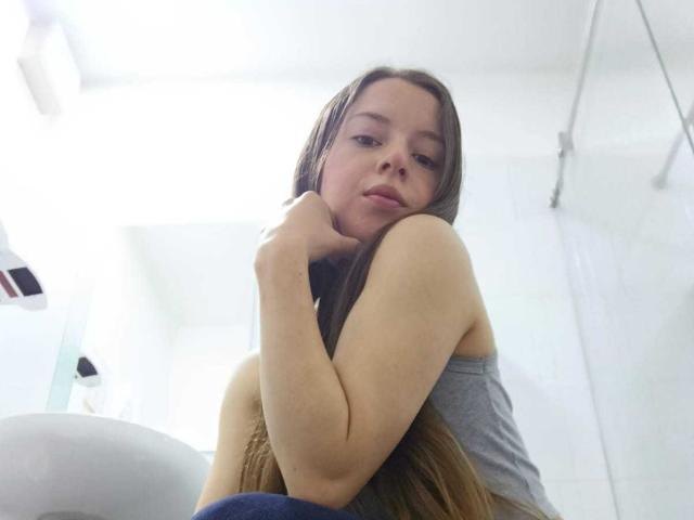 VioletaNova with long brown hair, wearing a grey tank top, hugging her knee, looking down with a slightly bored expression, in a bright, white-walled bathroom.