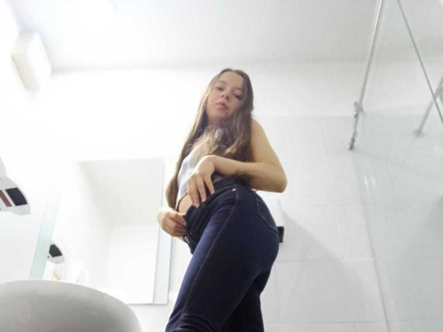 VioletaNova with long brown hair, wearing a white top and tight blue jeans, standing in a white-tiled bathroom, looking down at the camera with a slight smile.