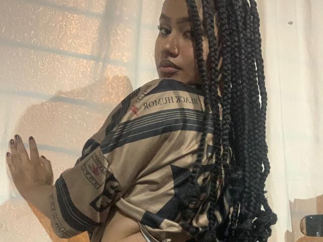 NoiraVale with long black braids, wearing a camo shirt, looking over her shoulder, showing a tattoo on her arm, and dark nail polish, in a softly lit room.